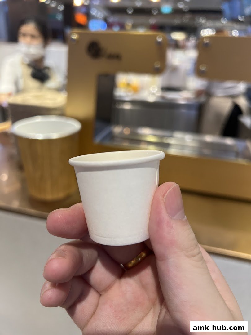 A hand holds a small white paper cup in front of a blurred beverage shop counter.