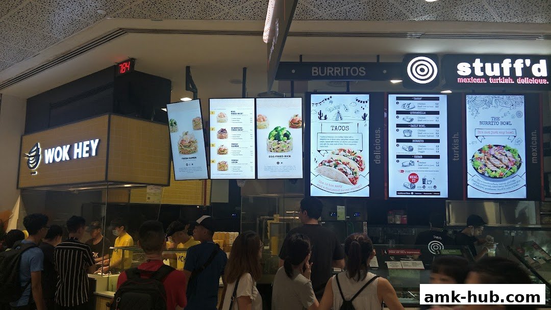A busy food stall with digital menus displaying various dishes and customers queuing.