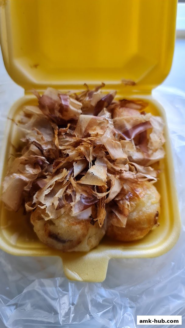 A close-up of takoyaki balls topped with bonito flakes in a yellow takeout container.