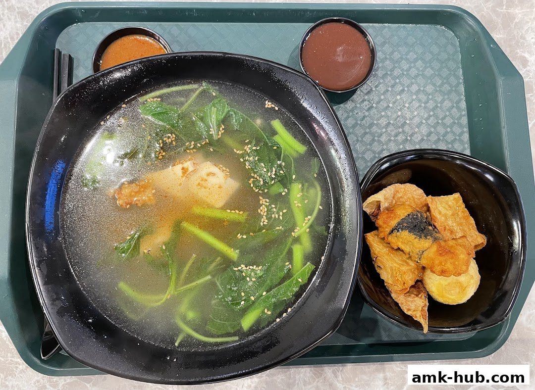 A bowl of Yong Tau Foo soup with vegetables and fried items, served with dipping sauces.