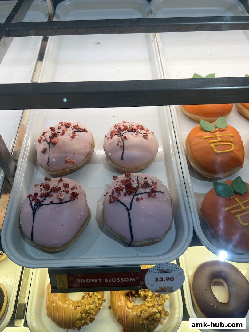 A display case filled with assorted Krispy Kreme donuts, featuring pink 'Snowy Blossom' and orange fruit-shaped varieties.