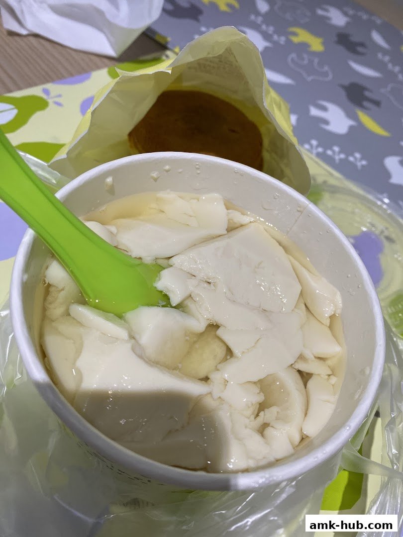 A close-up of a bowl of silky tofu pudding with a green spoon and a pastry in a bag.