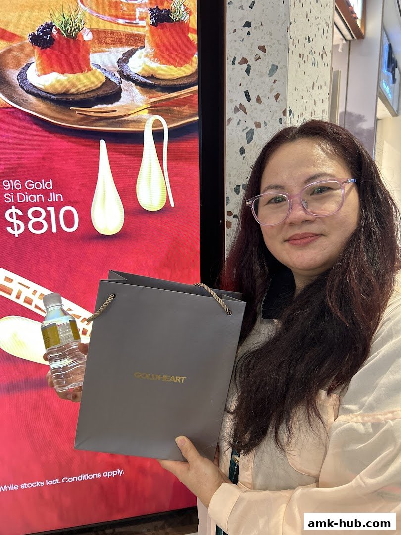 A woman holds a Goldheart shopping bag in front of a display featuring gold earrings and appetizers.