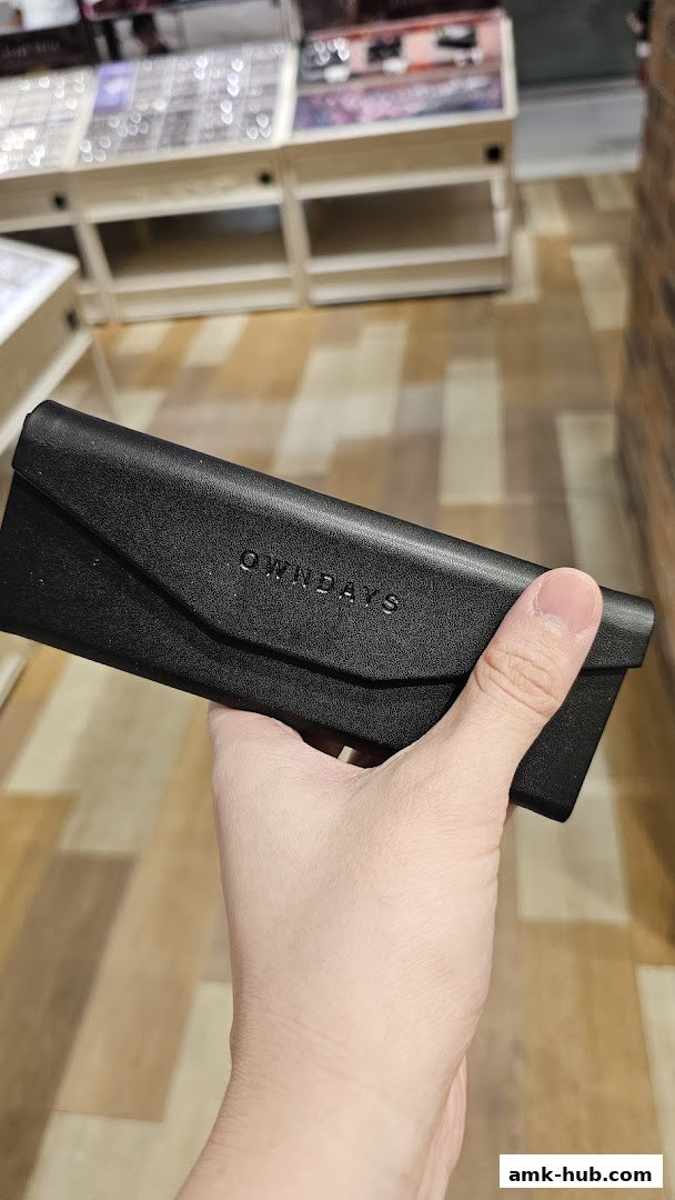 A hand holds a black OWNDAYS branded glasses case in front of a display of eyewear.