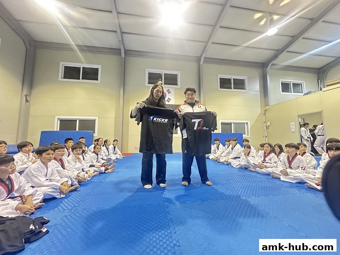 Two instructors hold up branded t-shirts to a seated audience of young Taekwondo students.