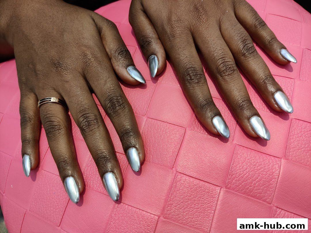 Close-up of hands with metallic silver almond-shaped nails on a pink textured surface.