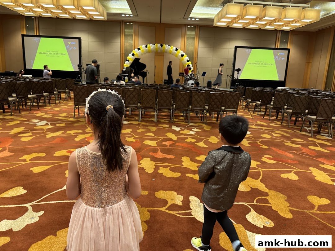 Two children stand in a hall with rows of chairs and a stage set up for an event.