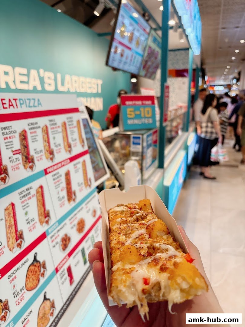 A person holds a slice of pizza in front of a brightly lit pizza shop counter.
