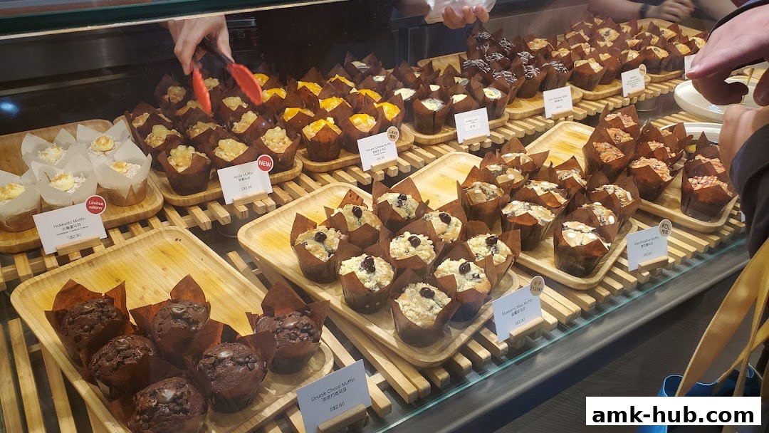 A display of various freshly baked muffins in a bakery, with customers selecting their choices.