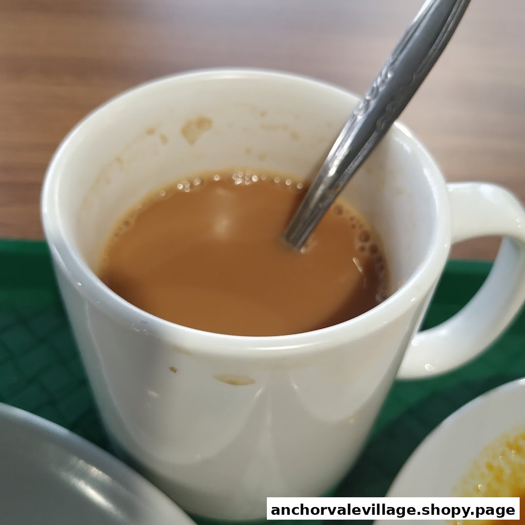 A close-up of a white mug filled with a frothy brown beverage, likely teh tarik, with a spoon.