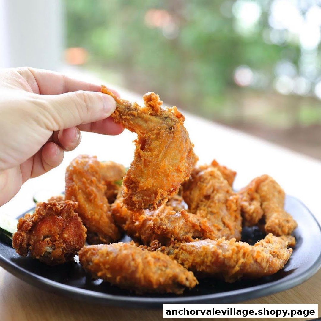 A hand holds a crispy fried chicken wing, with a plate full of golden-brown wings in the foreground.