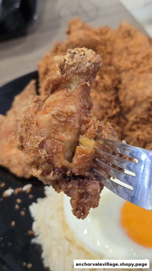 A close-up of a crispy fried chicken piece on a fork, with rice and a fried egg in the background.