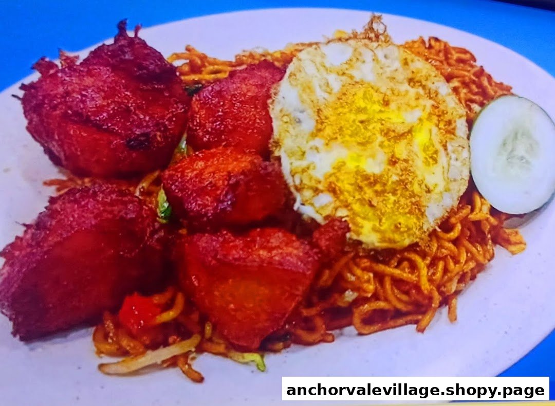 A plate of fried noodles with crispy chicken pieces, a fried egg, and a cucumber slice.