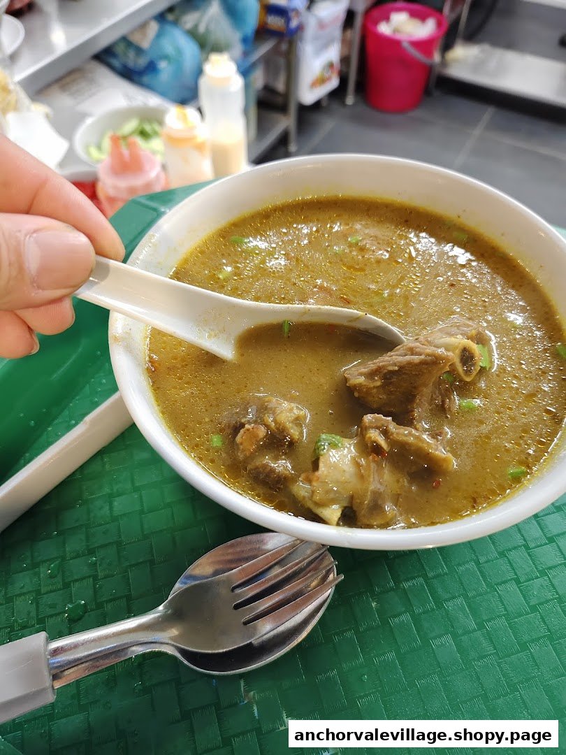 A hand holds a spoon in a bowl of rich mutton soup with meat chunks on a green tray.