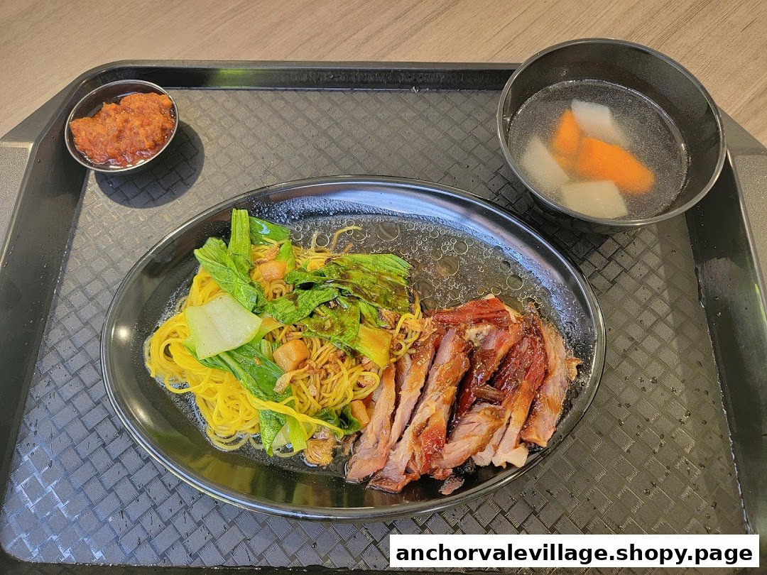 A delicious plate of char siew noodles with vegetables, served with chili and a clear soup.