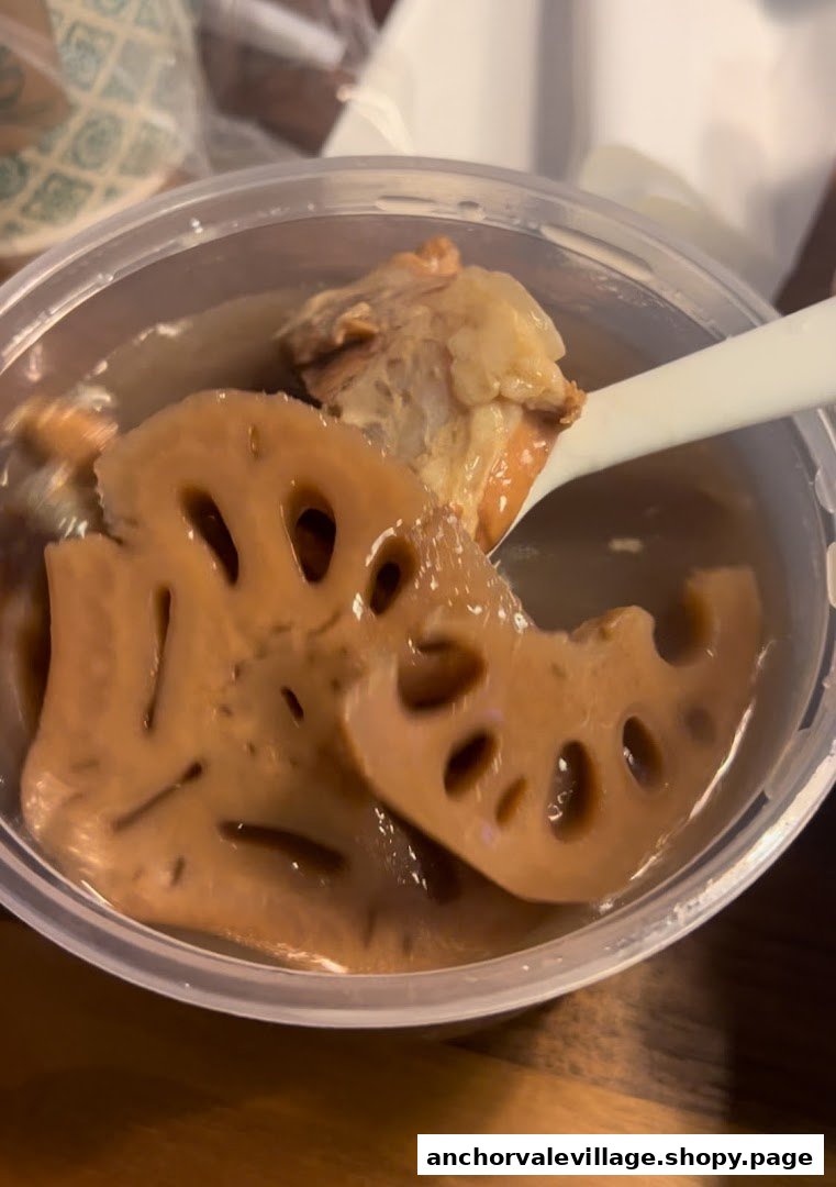 A close-up of a clear plastic container filled with lotus root soup and tender meat, with a white spoon.