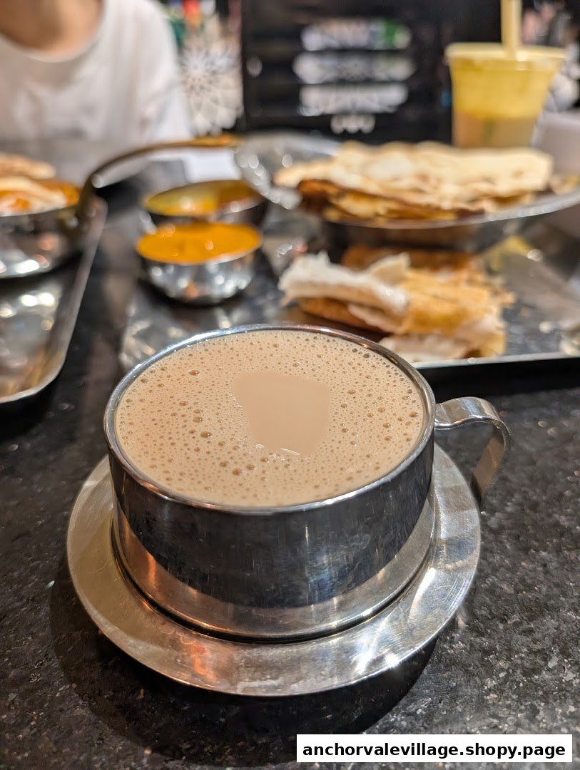 A close-up of a frothy beverage in a steel cup, with Indian food and condiments in the background.
