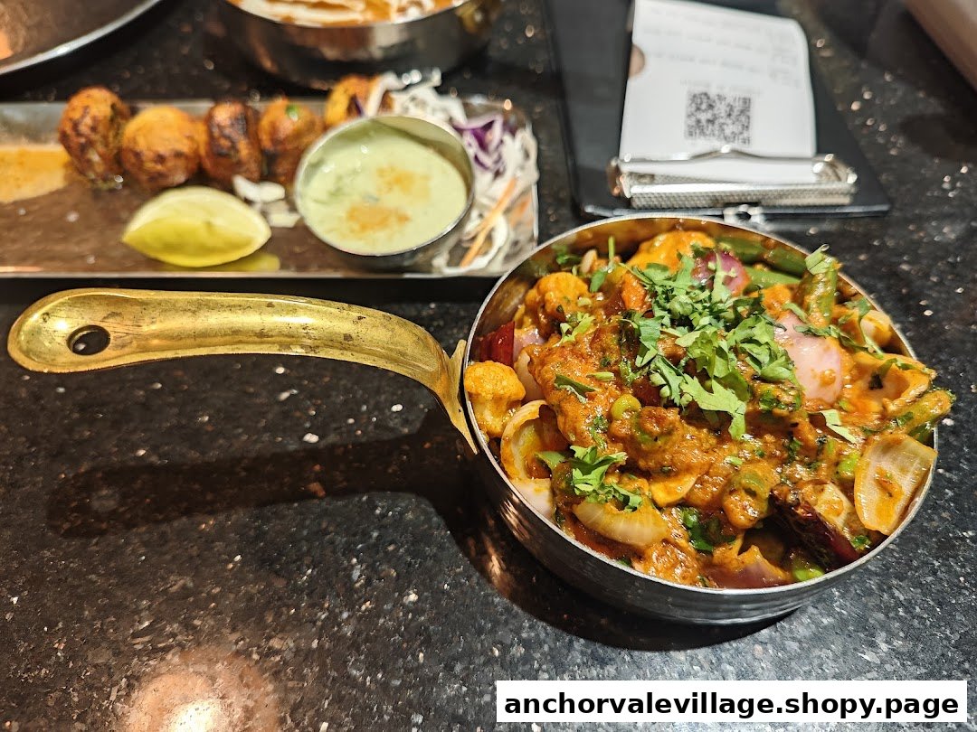 A vibrant vegetable curry in a brass-handled pot and appetizers with dipping sauce on a dark table.