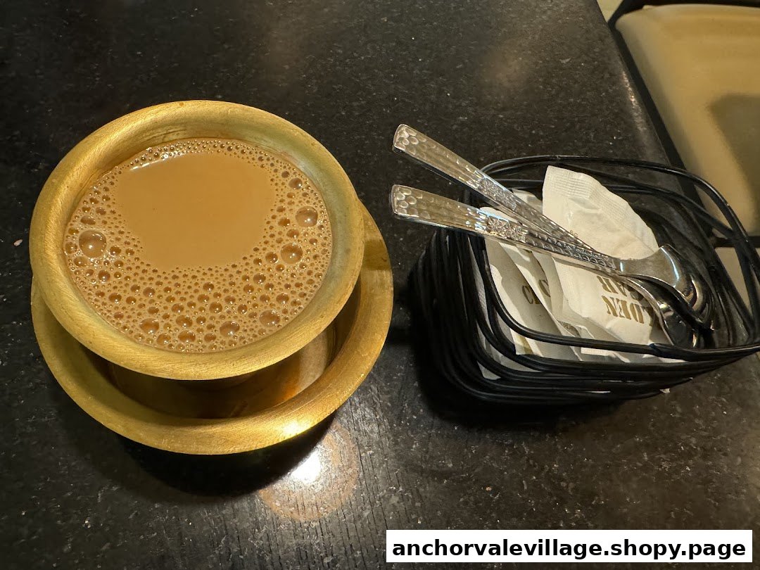 A warm cup of chai in a traditional brass cup, alongside a cutlery holder on a dark table.