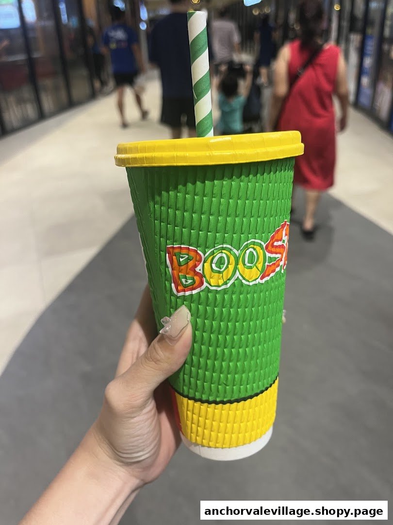 A hand holds a vibrant green Boost Juice cup with a striped straw in a mall setting.