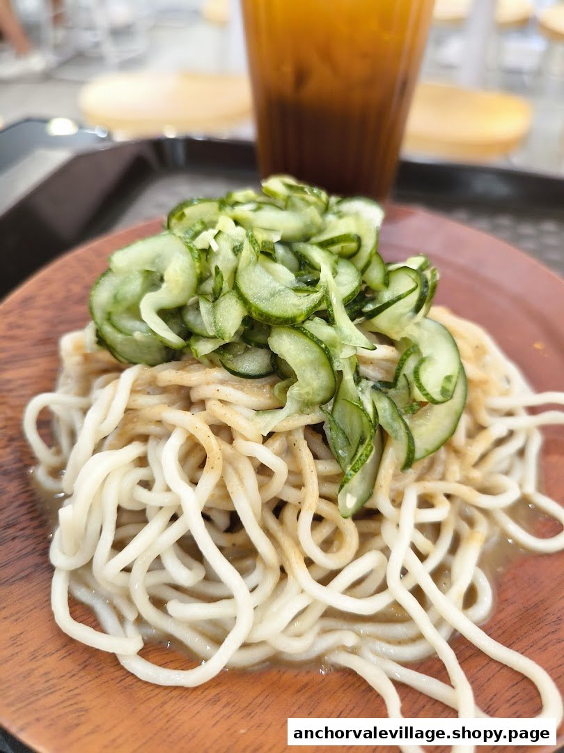 A close-up of cold noodles topped with sliced cucumbers on a wooden plate, with a drink in the background.