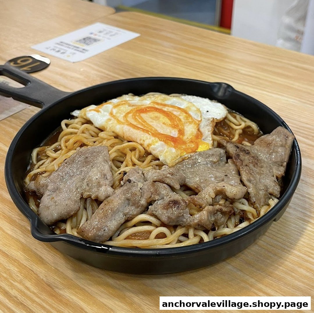 A close-up of noodles with sliced meat and a fried egg served in a black pan on a wooden table.
