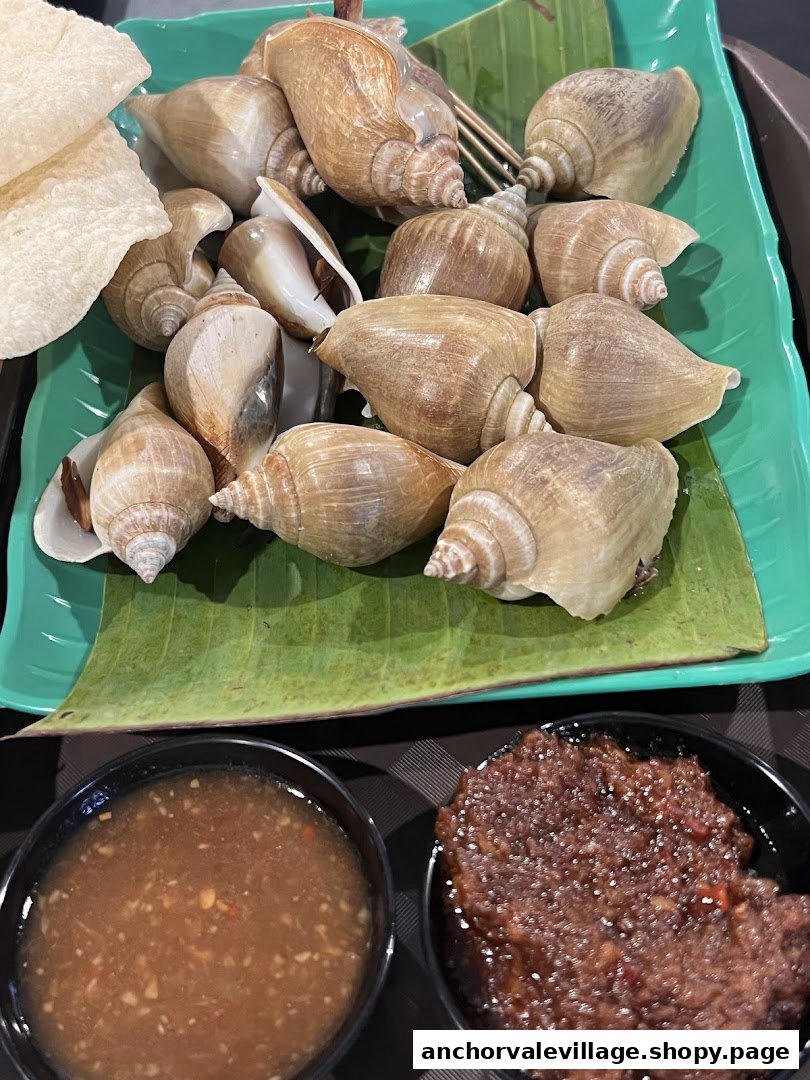 A plate of cooked sea snails with dipping sauces and crackers on a banana leaf.