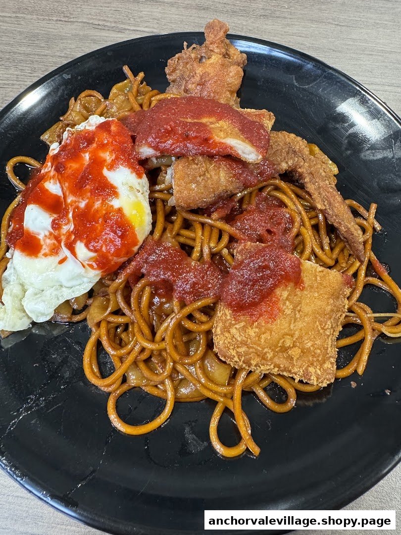 A plate of stir-fried noodles topped with fried chicken, fried egg, and chili sauce.