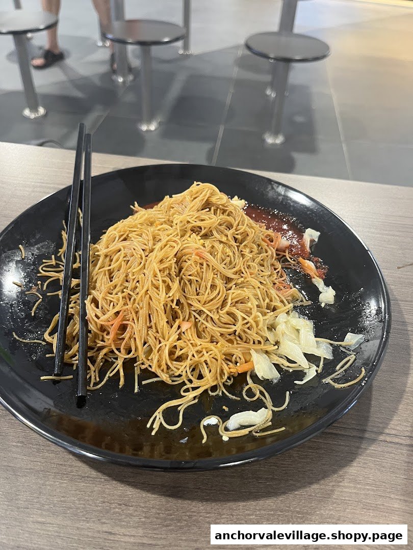 A plate of stir-fried noodles with chopsticks and chili sauce on a wooden table.