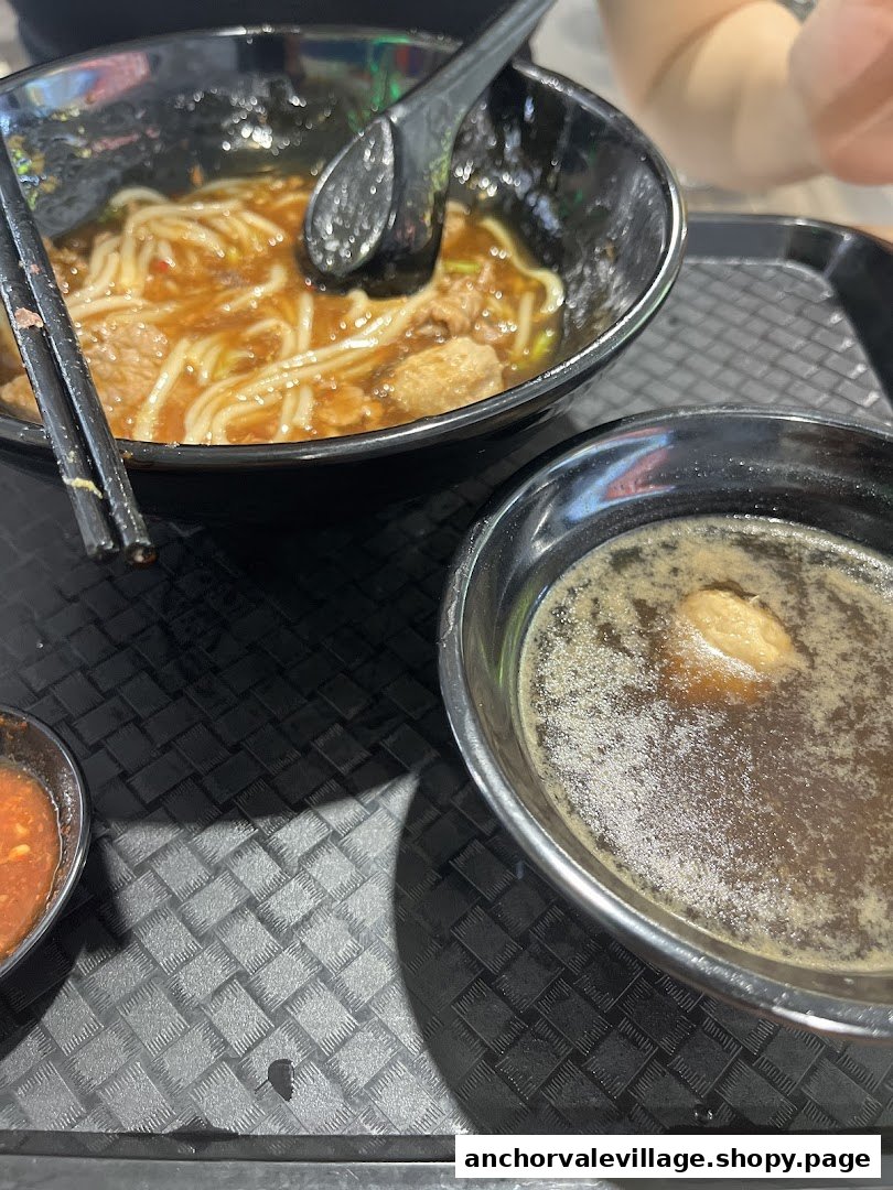 A close-up of a delicious bowl of Hainan beef noodles with a side of soup and chili sauce on a tray.