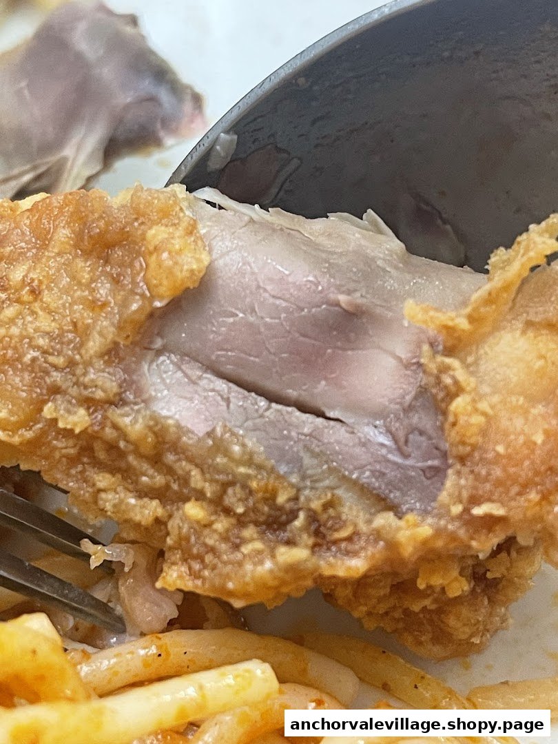 A close-up of a piece of fried chicken, revealing its juicy interior, with pasta and a utensil.