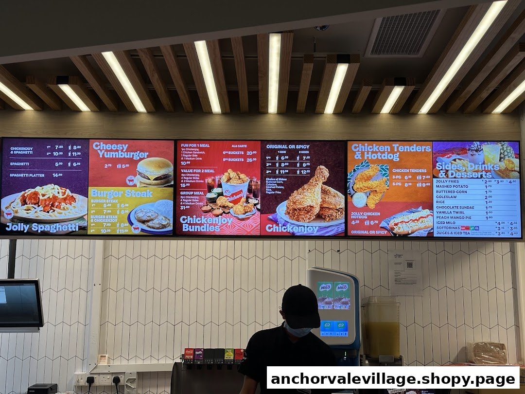 Digital menu boards displaying various food and drink items at Jollibee, with a staff member at the counter.