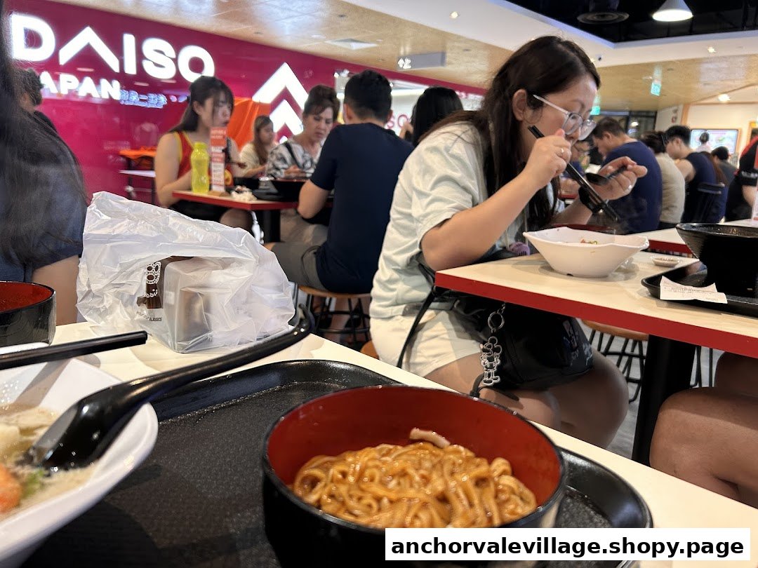 Diners enjoy meals in a bustling food court, with a bowl of noodles in the foreground.