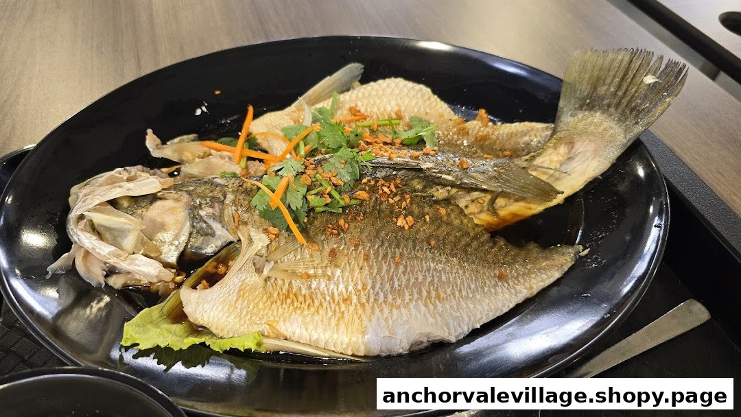 A beautifully presented steamed fish dish, garnished with fresh herbs and crispy fried garlic, on a black plate.