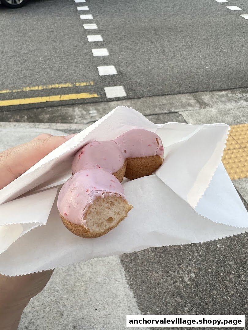 A hand holds three pink-glazed donuts in a white paper bag, with a street in the background.
