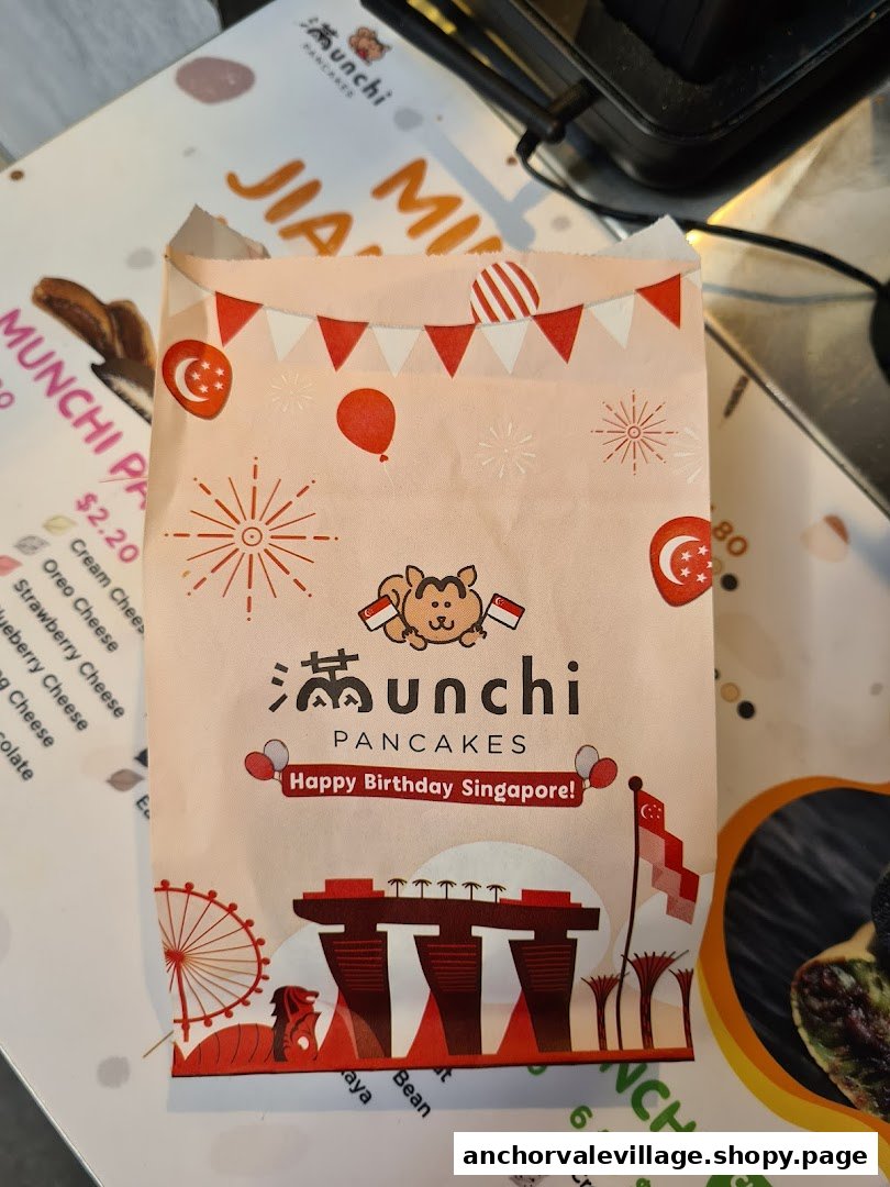 A festive Munchi Pancakes branded paper bag featuring Singaporean motifs, placed on a table with a partial menu.