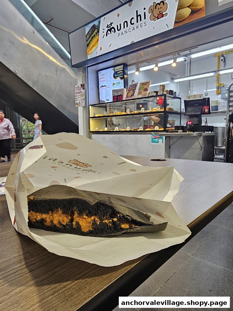 A charcoal pancake with vibrant orange filling in a paper bag, with the Munchi Pancakes shop visible behind.