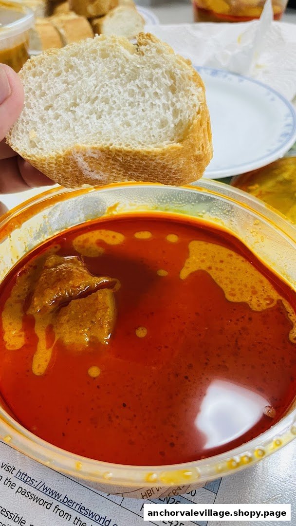 A hand holds a piece of bread, ready to dip into a rich, red curry with meat in a clear container.