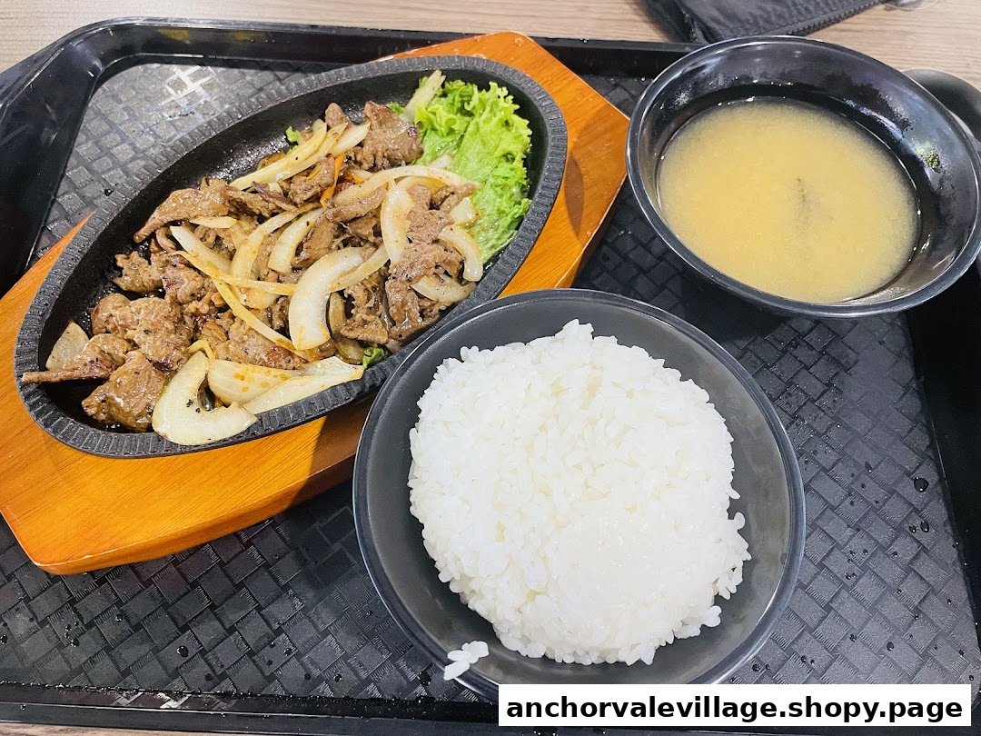 A sizzling plate of beef with onions, served with a bowl of white rice and a side of soup.
