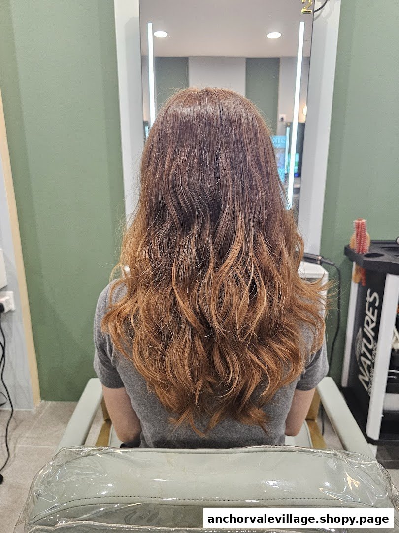 A person with long, wavy, reddish-brown hair sitting in a salon chair, facing a mirror.