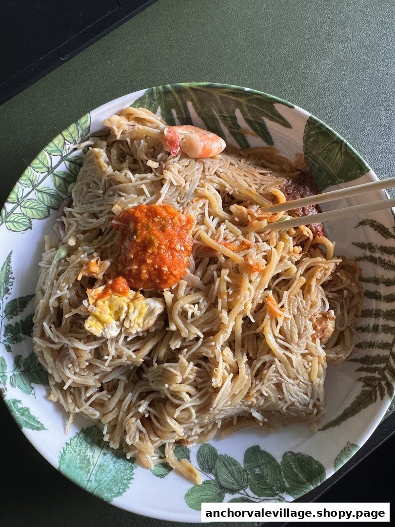 A close-up of a plate of Hokkien Mee with chili, shrimp, and egg, ready to be eaten with chopsticks.
