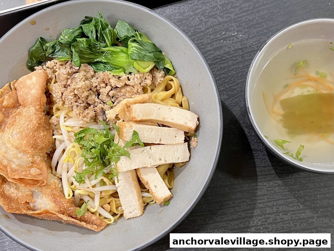 A vibrant bowl of vegan noodles with mock meat, vegetables, fried wontons, and a side of clear soup.