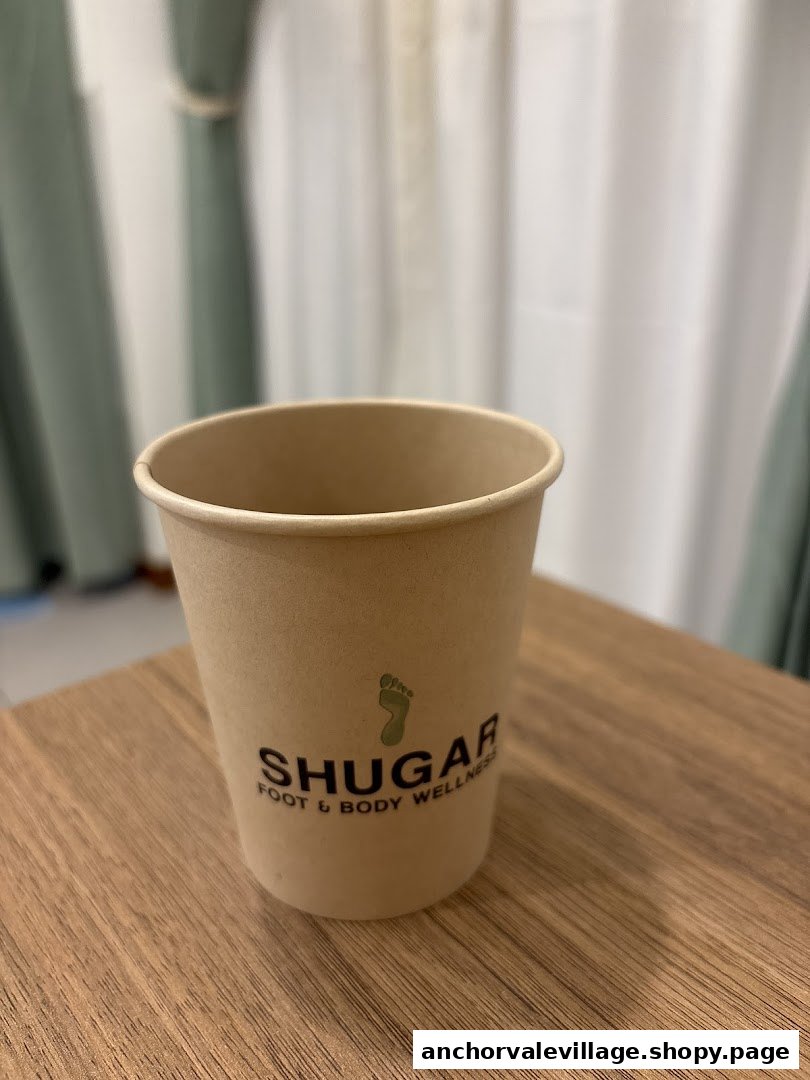 A branded paper cup from Shugar Foot & Body Wellness on a wooden table.