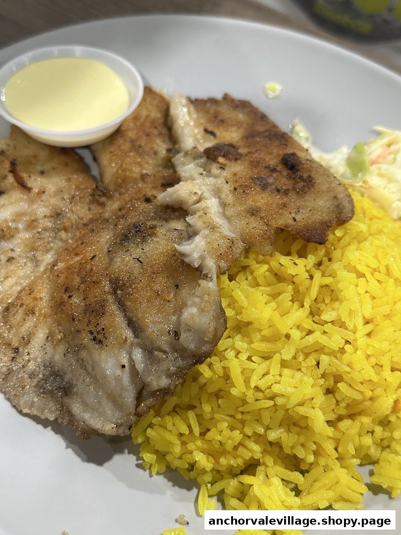 A close-up of a plate featuring pan-fried fish fillets, vibrant yellow rice, coleslaw, and a side of sauce.