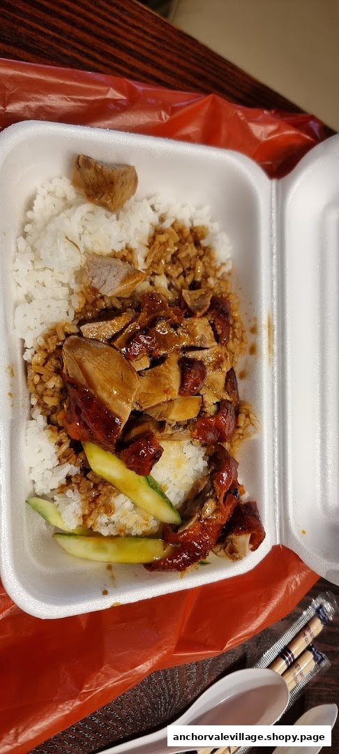 A takeaway box featuring roasted duck and char siu over rice, garnished with cucumber slices.