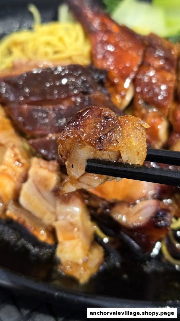 Close-up of glistening roasted pork belly held by chopsticks, with char siu and noodles in the background.