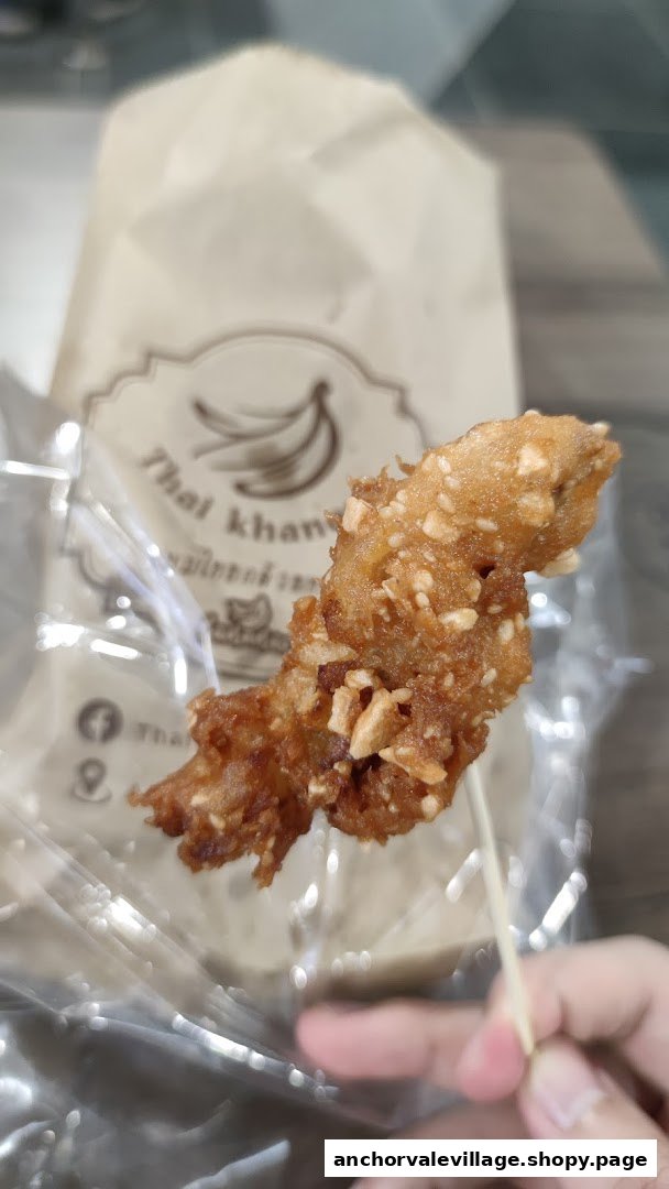A hand holds a crispy, peanut-crusted fried banana from Thai Khanom Bananas, with branded packaging visible.