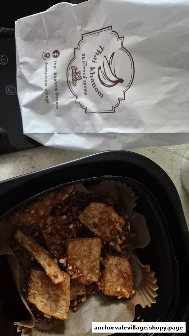 A container of crispy fried Thai banana snacks next to a branded 'Thai Khanom Bananas' paper bag.