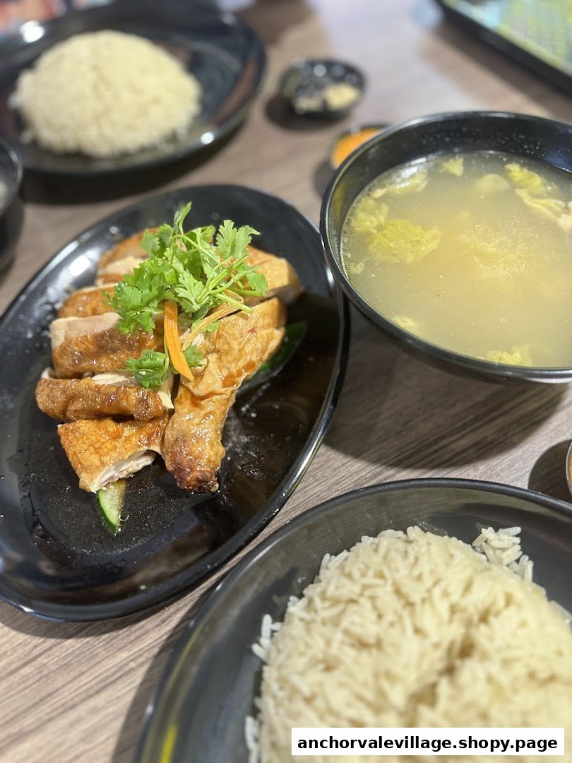 A close-up of a delicious chicken rice meal with soup and condiments on a wooden table.