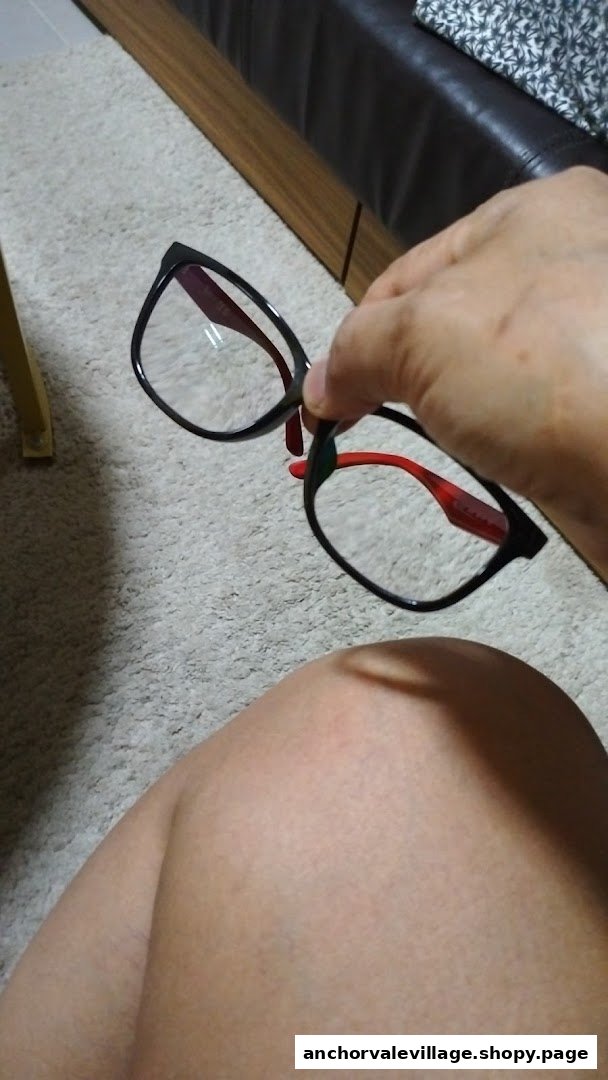 A hand holds a pair of black-framed eyeglasses with red temples, showcasing the product.